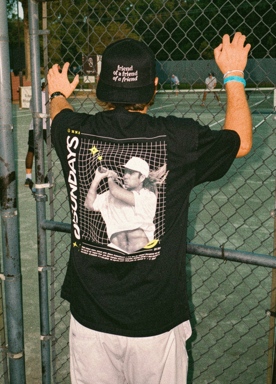 Person wearing a black t-shirt with a graphic design, standing behind a chain-link fence.